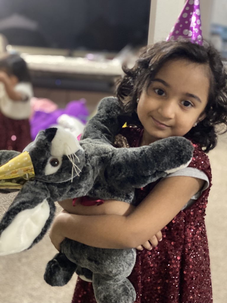 A young girl in a colorful party hat joyfully holds her beloved stuffed animal, radiating happiness.