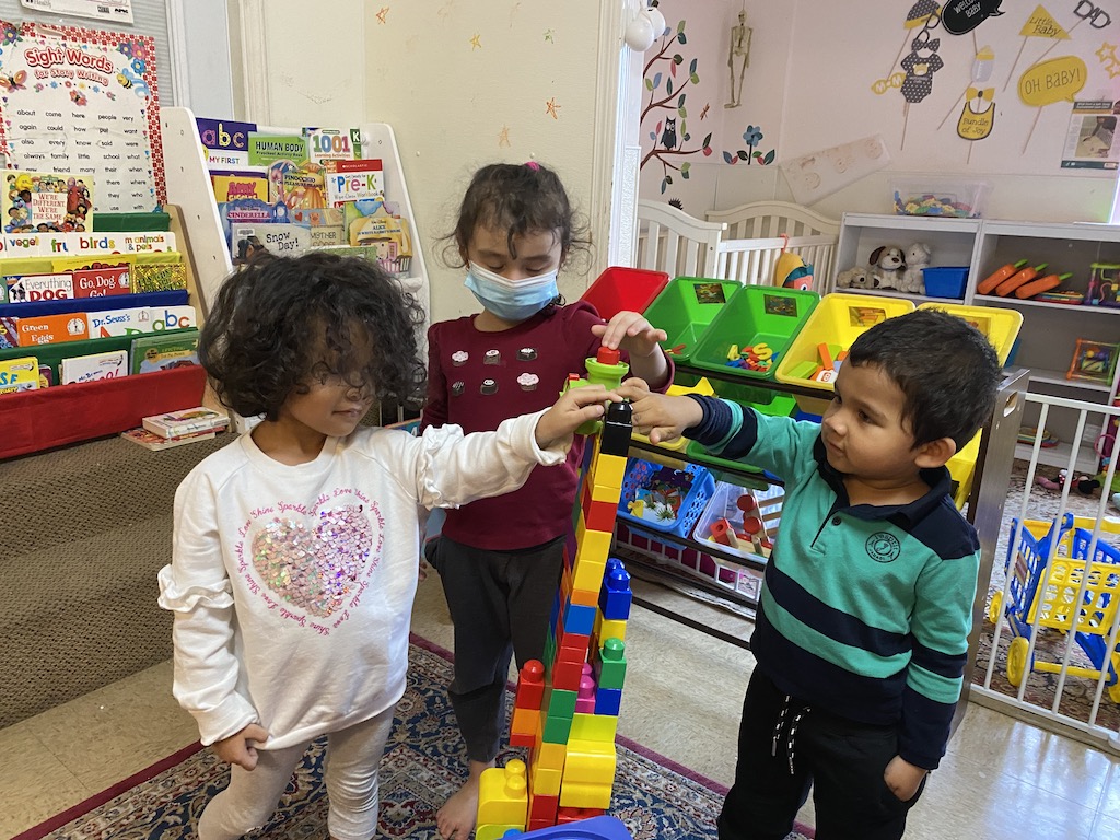 Three children engaged in play, building structures with colorful blocks in a bright classroom setting.